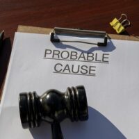 A probable cause document rests in a clipboard beside a gavel on a wooden desk, representing the documentation needed for court-authorized action.